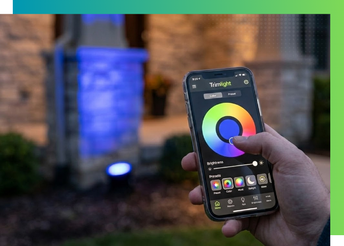 A hand holding a smartphone showing the Trimlight mobile app interface to change the color of a blue-tinted outdoor uplight.